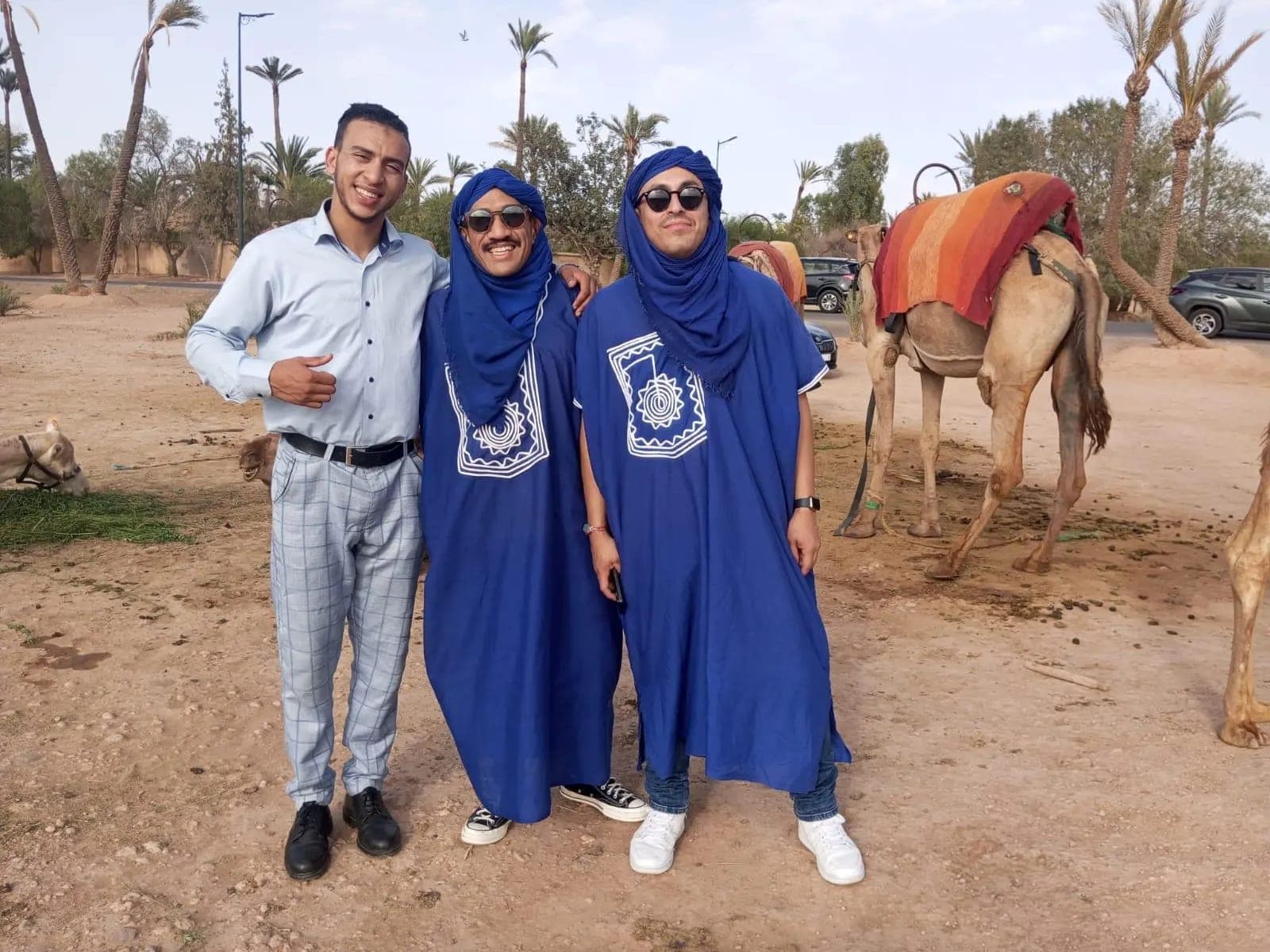 Two clients in blue Tuareg dress with their chauffeur in the Marrakech palm grove, a saddled dromedary behind them