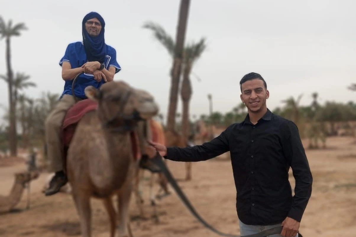 A client riding a dromedary in the Marrakech palm grove, accompanied by a guide holding the lead rope